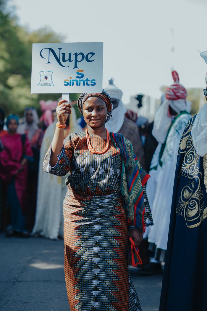 A woman in traditional Nupe attire holds a sign during a cultural parade showcasing rich African heritage.